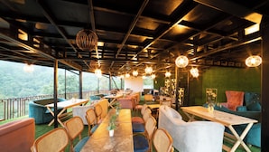 An indoor seating area with tables, chairs, and sofas, featuring a view through large windows. - Mudras Grove Resort (Nainital)