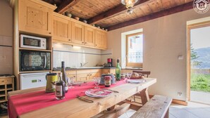 Una mesa de comedor de madera dispuesta con platos, vasos y una botella, en una cocina con armarios de madera y vistas al exterior.