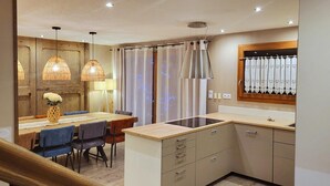 Una cocina moderna con una isla central, suelo de madera, luces colgantes y una zona de comedor con sillas azules y una mesa de madera.