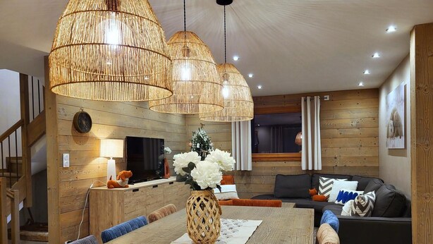 Una sala de estar moderna con piso de madera, una mesa de comedor con camino de mesa decorativo, un sofá con cojines y un televisor en la pared.