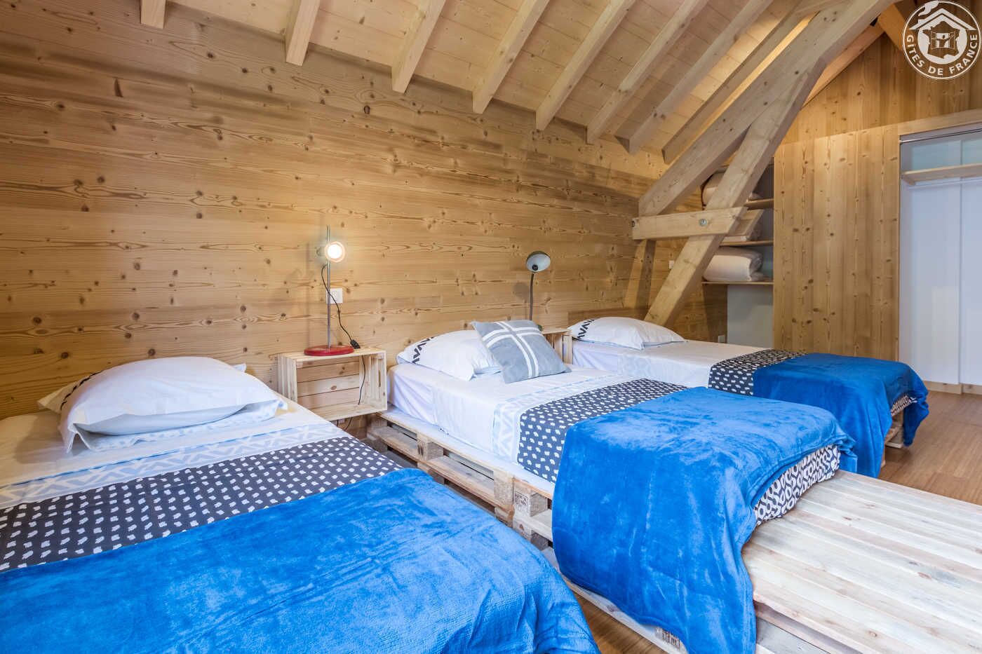 A wooden cabin with three beds, each with a blue blanket, and a nightstand with a lamp.