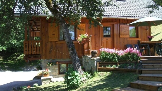 Una cabaña de madera con zócalo de piedra, un porche con mobiliario exterior y un jardín con flores y plantas.