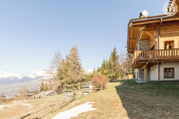 A wooden chalet with a balcony, stone walls, and a view of snow-capped mountains.