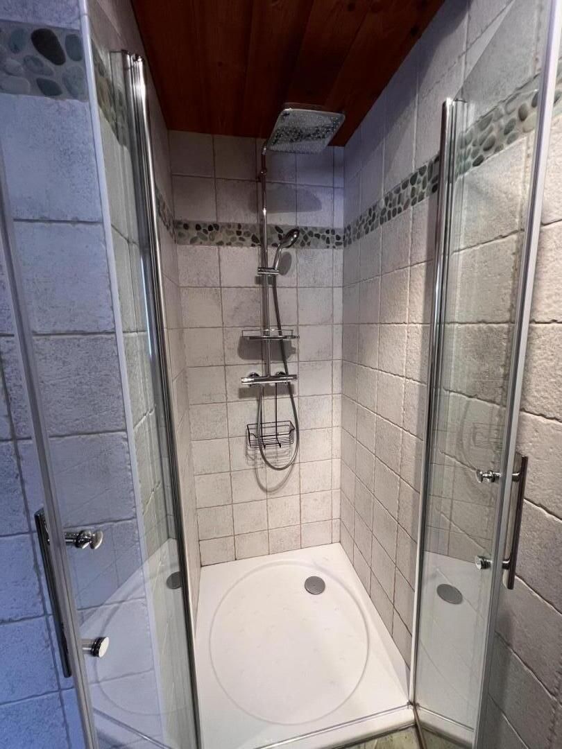 A glass-enclosed shower stall with a white bathtub, tiled walls, and a wooden ceiling.