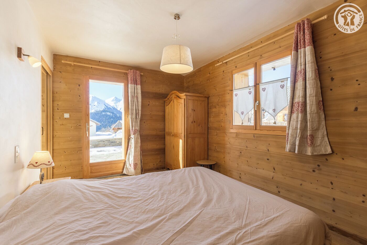 A wooden room with a bed, a window with curtains, a lamp, and a wardrobe.