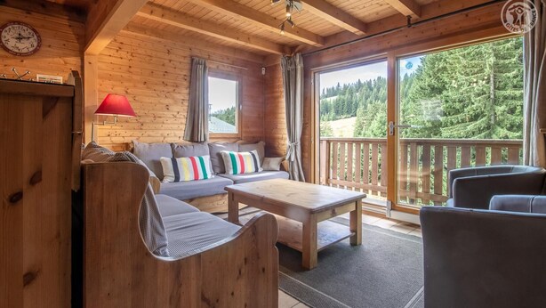 A cozy wooden cabin interior with a sofa, chairs, a table, and a large window offering a forest view. - LE Chomesi - Hauteluce-les-saisies (HAUTELUCE LES SAISIES)