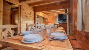 A wooden dining table set with plates, glasses, and cutlery, with a television and kitchen visible in the background. - LE Chomesi - Hauteluce-les-saisies (HAUTELUCE LES SAISIES)