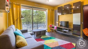 Una sala de estar moderna con sofá, un tapete colorido, un televisor y un balcón con cortinas amarillas.