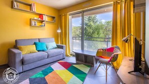 Un salón con un sofá gris, un tapete colorido y un sillón con cojín de parches. Se ven estanterías y una ventana con cortinas.