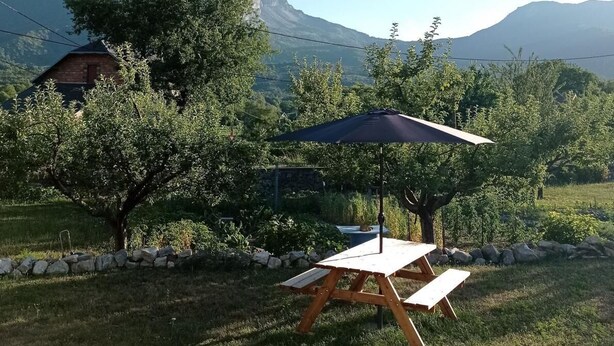Ein Picknicktisch mit Sonnensegel in einer grünen Umgebung mit Bäumen und einem Berg im Hintergrund.