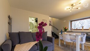 Ein modernes Wohnzimmer mit einem grauen Sofa, einem Esstisch mit Stühlen, einer Topfpflanze und Blick auf eine Küche und eine Treppe.