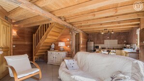 A cozy wooden cabin interior with a sofa, a chair, a kitchen area, and a staircase.