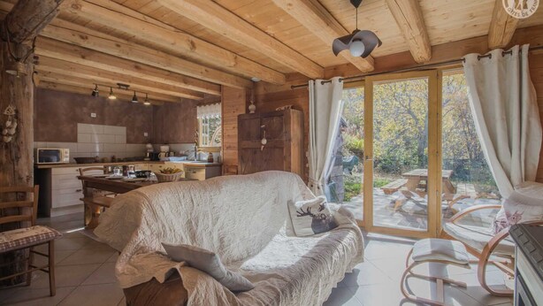 A cozy cabin interior with a covered sofa, wooden ceiling beams, a kitchen area, and a sliding glass door leading to an outdoor patio.