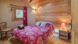 A cozy bedroom with a wooden ceiling, a bed with a red patterned bedspread, a small bedside table with a lamp, and a window with a curtain.