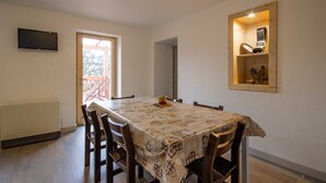 Un área de comedor con una mesa dispuesta para cuatro personas, un televisor montado en la pared y un estante de madera con adornos.
