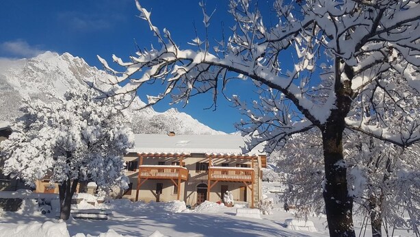 Un edificio cubierto de nieve con un balcón de madera, rodeado de árboles cargados de nieve y montañas al fondo.
