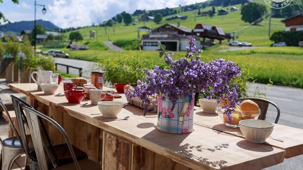 Una mesa de madera con varios cuencos y un jarrón de flores moradas, frente a un paisaje con campos verdes y casas.