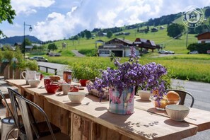 A wooden table with various bowls and a vase of purple flowers, set against a scenic backdrop of green fields and houses.