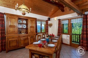 Uma sala de jantar de madeira com uma mesa posta para quatro pessoas, rodeada por cadeiras. Há um armário de madeira com louças e um candelabro sobre a mesa.