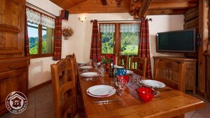 Une salle à manger en bois avec une longue table dressée pour quatre personnes, un téléviseur et une vue sur l’extérieur.
