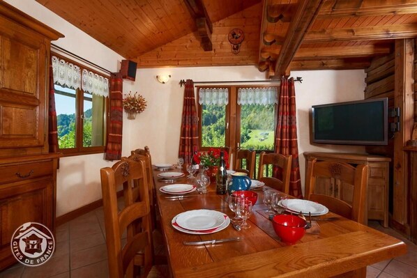 A wooden dining room with a long table set for four, a television, and a view of the outdoors.