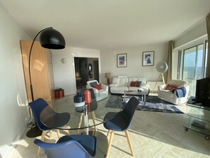 Ein modernes Wohnzimmer mit einem Glastisch, blauen Stühlen, einem weißen Sofa mit roten und blauen Kissen und einem großen Fenster mit Blick auf die Stadt.