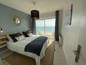 Ein Schlafzimmer mit Bett, großem Fenster, Meerblick, rundem Spiegel und einem Bild an der Wand.