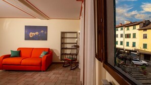 A room with a red sofa, a wooden rocking chair, a bookshelf, and a window overlooking a street with buildings and parked cars.
