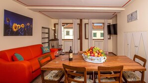 A dining area with a wooden table, four chairs, a fruit bowl, wine glasses, and a bottle. A red sofa and a painting of sunflowers are on the left.