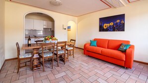 A dining area with a wooden table and chairs, a kitchen in the background, and a bright orange sofa with decorative pillows.
