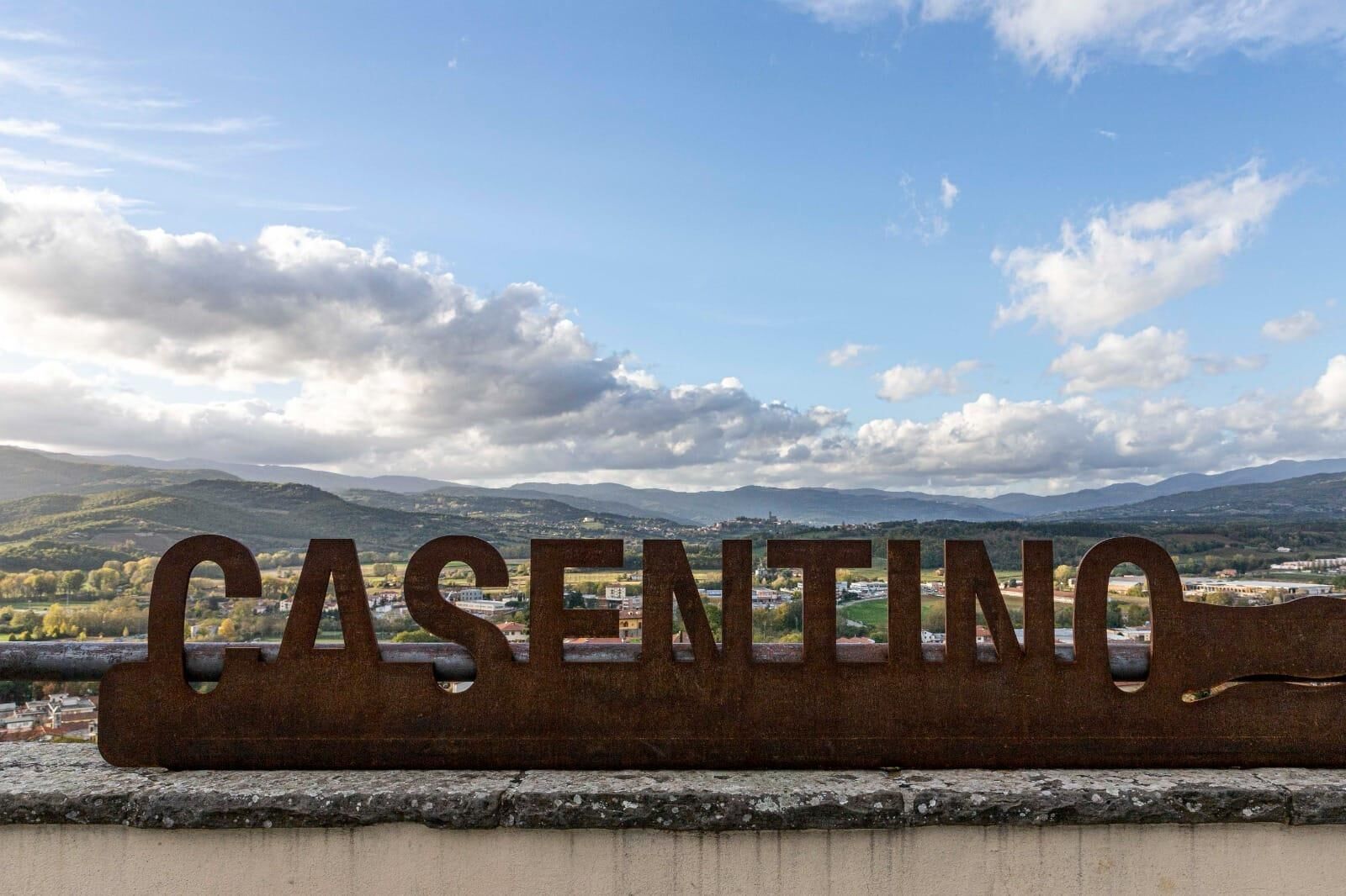 A metal sign with the word "CASENTINO" against a backdrop of mountains and a partly cloudy sky.