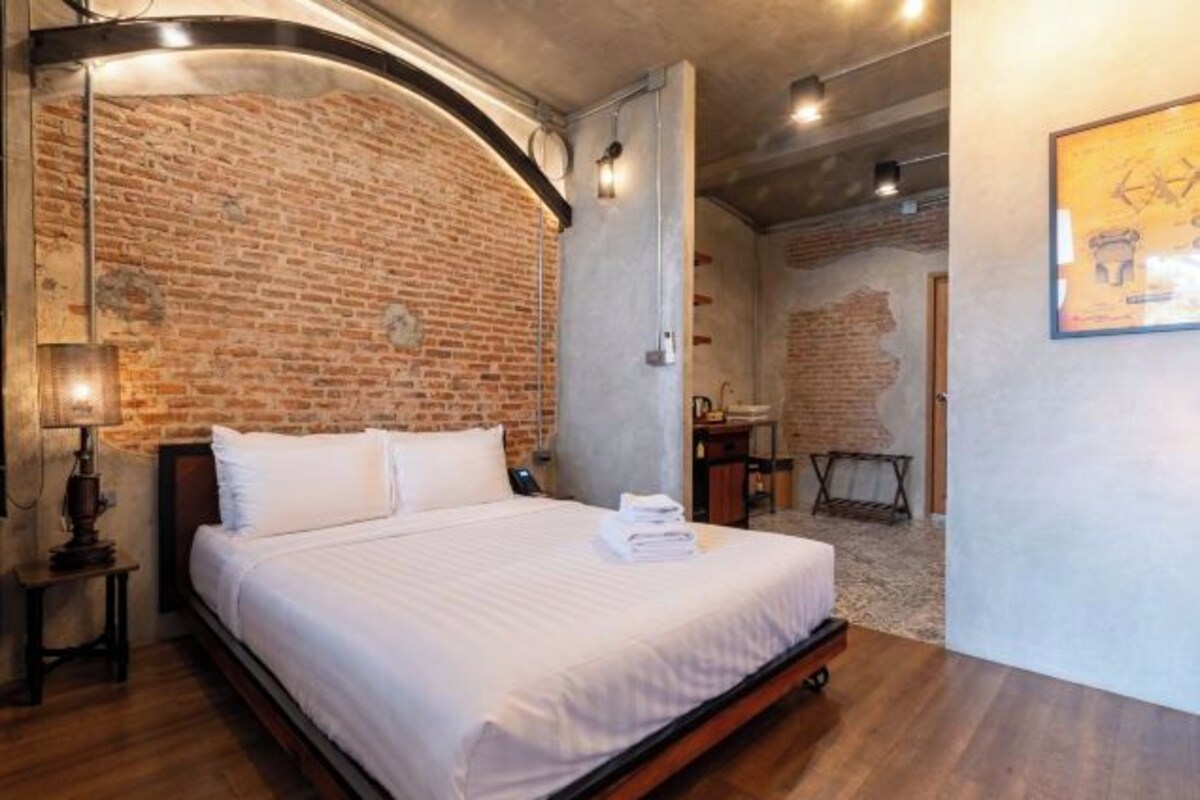 A room with a brick wall, a bed with white bedding, a desk, and a chair.