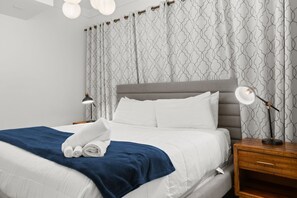A neatly made bed with white linens and a blue blanket, a bedside lamp, and a wooden nightstand with a drawer.