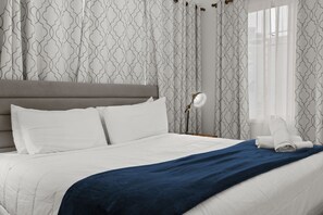 A neatly made bed with white and blue bedding, a grey headboard, and patterned curtains.