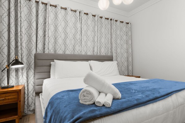 A neatly made bed with white linens and a blue blanket, a bedside lamp, and a patterned curtain.