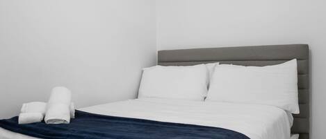 A neatly made bed with white linens and a dark blue blanket, a grey upholstered headboard, and a white pillow on the right side.