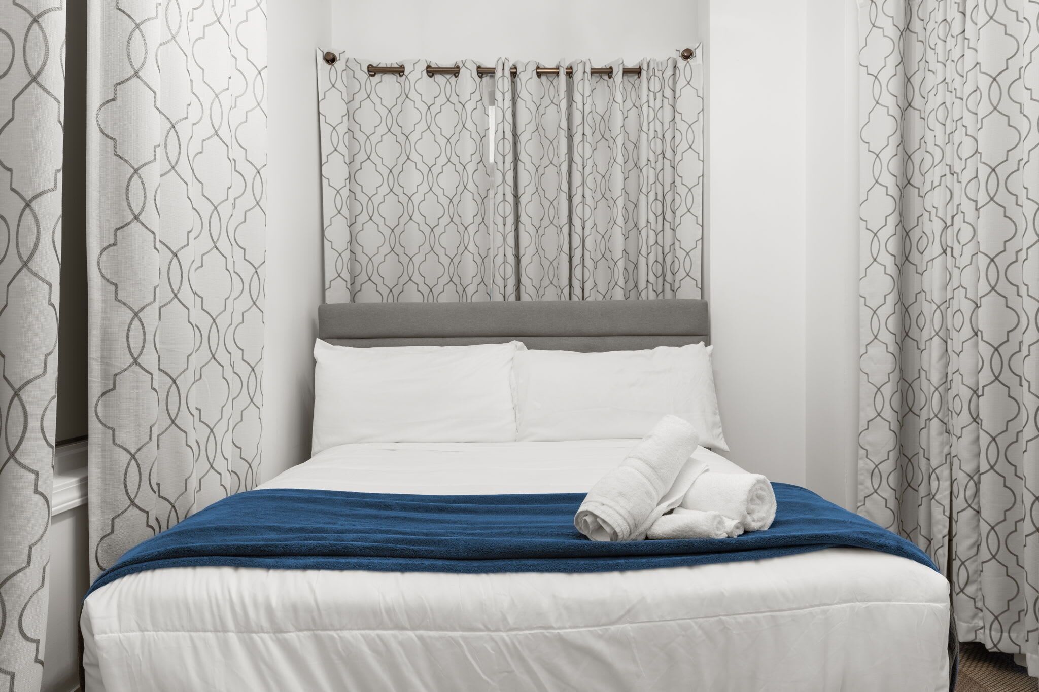 A neatly made bed with white bedding and a blue accent, a grey headboard, and patterned curtains.