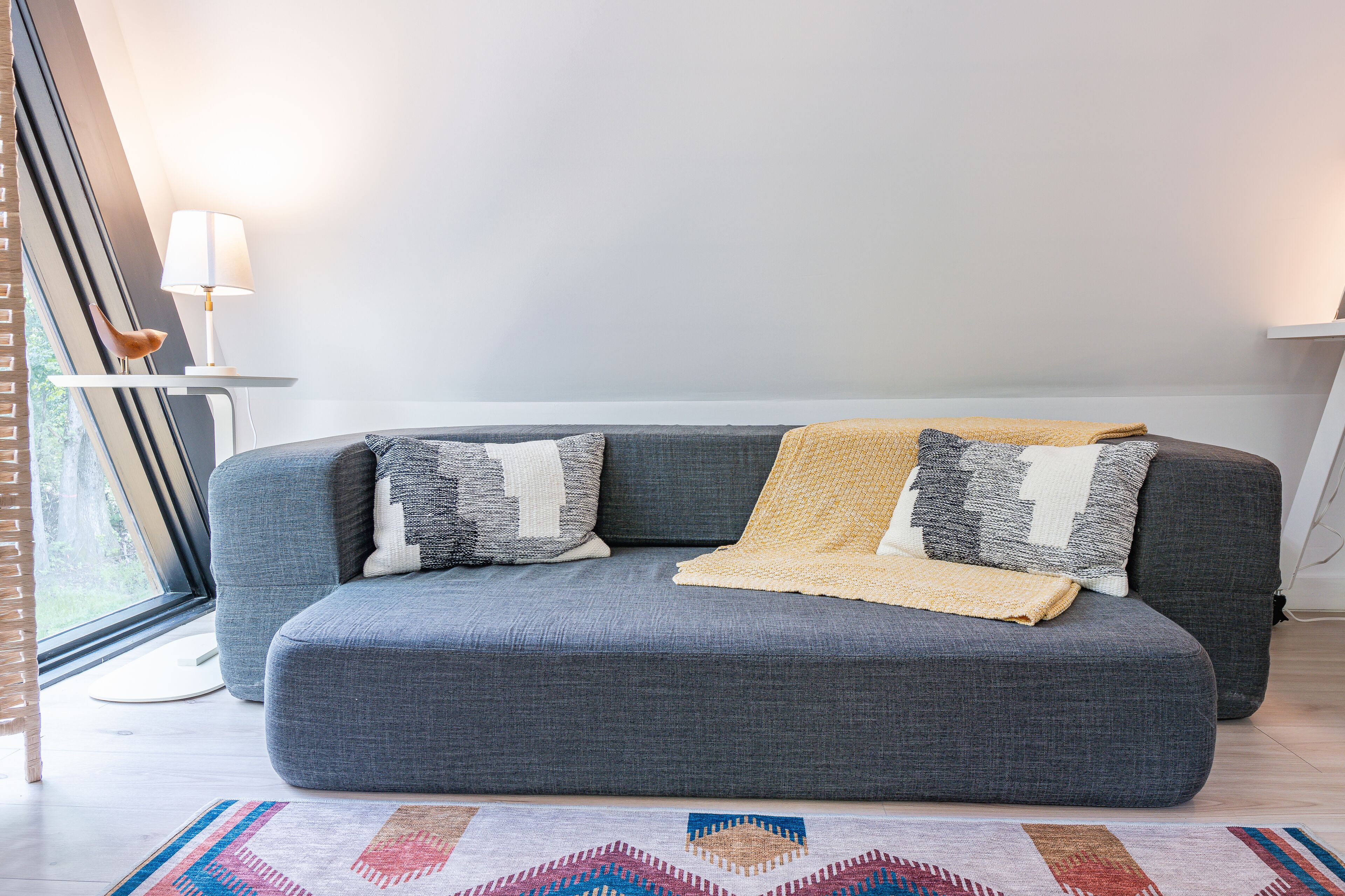 Un canapé gris agrémenté de coussins à motifs et recouvert d’un plaid en tissu, placé devant une fenêtre sur laquelle repose une lampe blanche, posée sur une petite table d’appoint.