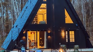 Una cabaña de madera triangular con un amplio ventanal, una fogata y una mesa en el exterior.