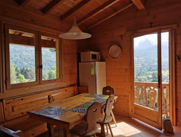 A wooden cabin with a dining table and chairs, a refrigerator, and a view of a mountainous landscape through large windows.