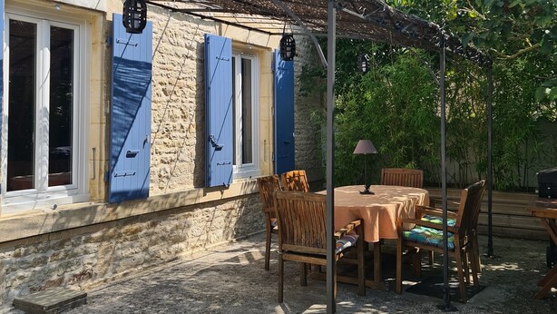 Een stenen gebouw met blauwe luiken, een overdekt terras met tafel en stoelen, en een tuin.