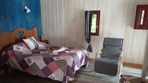 A bedroom with a wooden bed, a grey armchair, and a window with curtains. - Joli Pavillon Individuel et Privatif (Rigaud)
