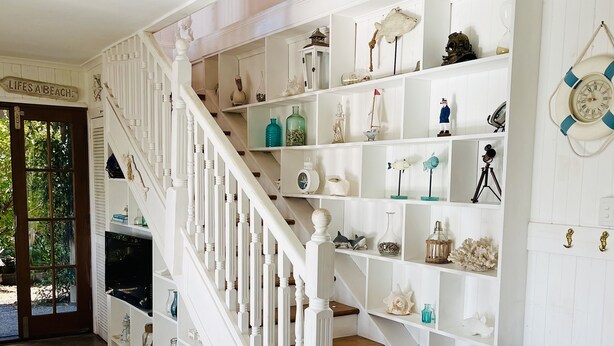 A staircase with white railings and wooden steps, leading to a landing with white shelves displaying various decorative items. - Coastal Queenslander- 2 minute walk to the beach! (Woodgate)