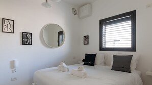 A neatly made bed with white linens and rolled towels, a round mirror, and a window with blinds. - Charming & Quiet 1 BR in Heart Tel Aviv SAFE ROOM (Tel Aviv-Yafo)