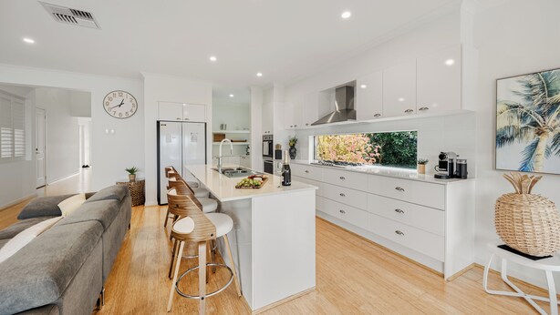 A modern kitchen with white cabinetry, a central island with bar stools, a large window with a view, and a dining area with a grey sofa.