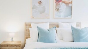 A neatly made bed with white and light blue bedding, a wooden nightstand with a lamp, and two framed pictures on the wall.
