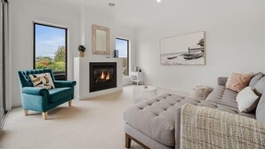 A modern living room with a fireplace, a teal armchair, a grey sofa, and a large window offering a view of the outdoors.