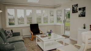 Ein modernes Wohnzimmer mit einem grünen Sofa, einem weißen Couchtisch und einem schwarzen Sessel. Große Fenster mit weißen Fensterläden, eine Heizung und ein kleiner Topf mit Pflanze auf dem Couchtisch.