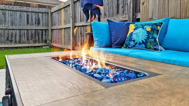 Een houten terras met een vuurkorf in het midden, omringd door een blauwe bank met bloemenkussens.