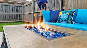A wooden deck with a fire pit in the center, surrounded by a blue sofa with floral cushions.